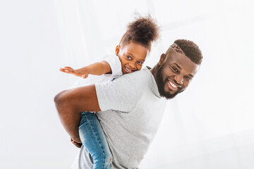 A happy African American father gives his young daughter a piggyback ride indoors. The father is...