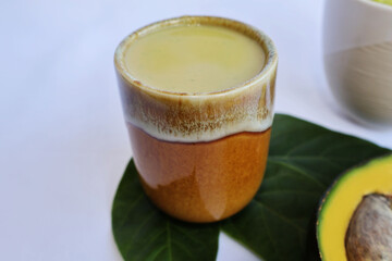 Avocado juice blended in two brown-yellow ceramic glasses. Placed on a green leaf on a white background

