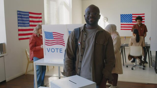 Portrait of African American man putting ballot in voting box and posing for camera with smile at polling place