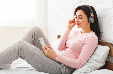 A woman is seated on a bed, listening to music through headphones, using smartphone. The room is...