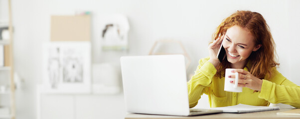 A woman sits at her desk in a modern office, talking on her cell phone, smiling, and holding a mug....