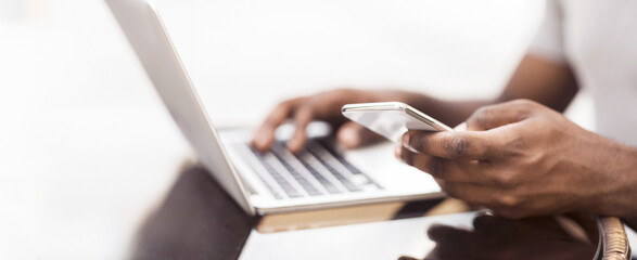 Business and technology concept. Millennial african american man using cellphone while working on laptop, panorama