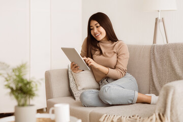 A woman sits on a couch at home, using a tablet. She is smiling and appears to be relaxed.