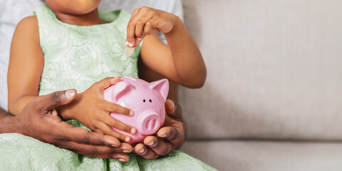 A young girl in a green dress is putting a coin into a pink piggy bank. An adult is helping her...