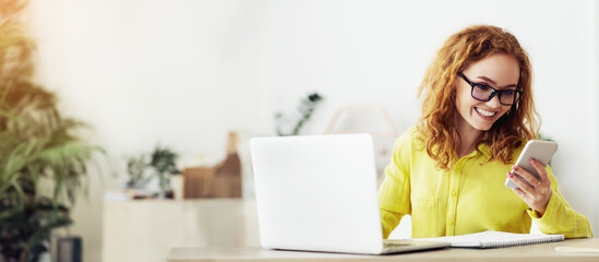 A woman with red hair and glasses smiles while looking at her smartphone. She is sitting at a desk...