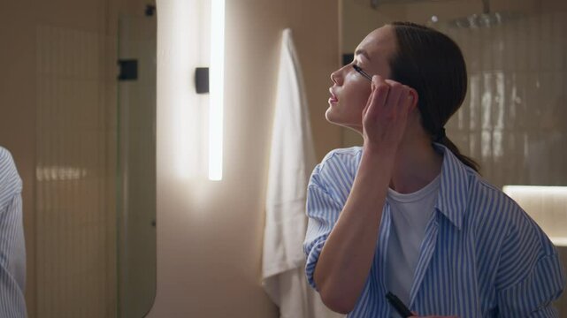 Relaxed girl applying mascara in bathroom mirror. Smiling woman admiring beauty