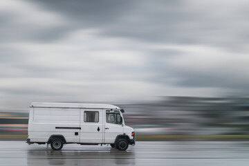 Old white cargo van is driving down the street in the rain with a motion blur background