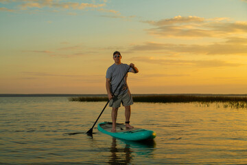 A man in shorts and a T-shirt swims on a lake on a stand-up paddle board at sunset