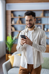 Vertical shot of a young Indian man at home, standing in a room and smiling while using a mobile...