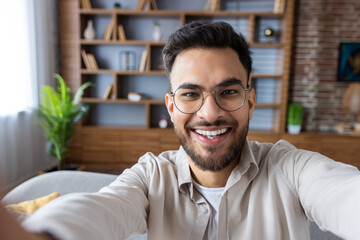 Close-up photo of young smiling Indian man in glasses and beard at home, holding camera and talking on video call