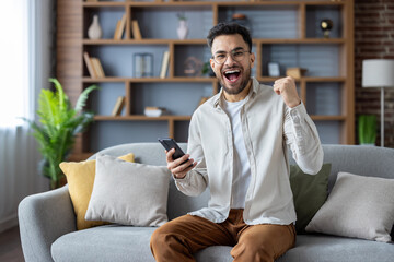 Portrait of happy young Indian man rejoicing at home sitting on sofa with achievement and success...