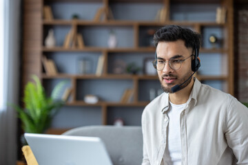 Obraz premium Indian young man wearing a headset is sitting on the sofa at home and talking remotely on a video call through a laptop