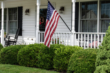 American flag flying from front porch of white colonial home with black shutters and rocking chair