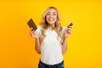 Naklejka premium This image features a young woman with long blonde hair smiling widely while holding two chocolate bars in her hands. She is standing in front of a bright yellow background.