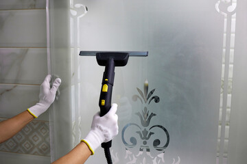 A woman in white gloves using a professional steam cleaner washes the glass door of the shower cabin.