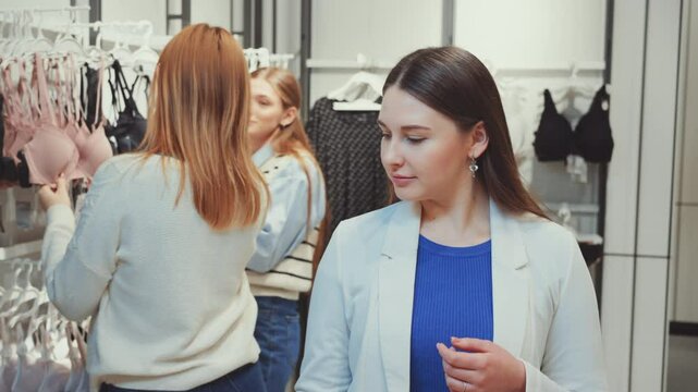 Three women shopping for lingerie, examining different bras and undergarments in store. Females focused on selecting items, showcasing variety of choices. Concept of fashion and retail therapy