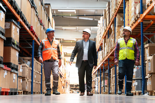 Manager and factory employees work together to warehouse aisle. Three men they are wearing hard hats and reflective vests.