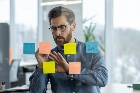 Close-up photo of a serious and focused young man working in an office, standing near a transparent information board and making notes on colored sticky notes