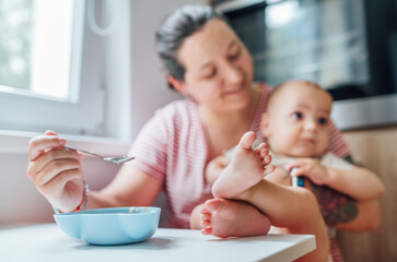 Mother lovingly feeds her baby in cozy kitchen showcasing moment of tender care and bonding. Baby, with feet playfully extended, looks content and curious, adding touch of warmth and intimacy to scene