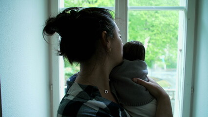 Mother holding newborn baby by a window, both looking outside, capturing a quiet moment of bonding and reflection