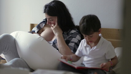 Mother breastfeeding newborn baby while older son reads a book beside her. The mother, in a plaid shirt, nurtures her baby, capturing a quiet moment of family life and sibling relationships on the bed