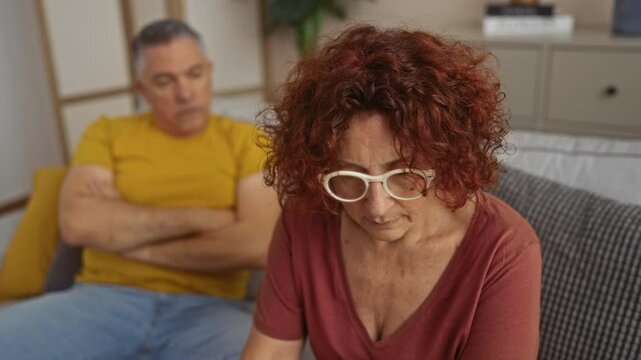 Middle-aged couple sitting indoors in a living room, looking upset and avoiding eye contact with each other, highlighting frustration and tension in their relationship.
