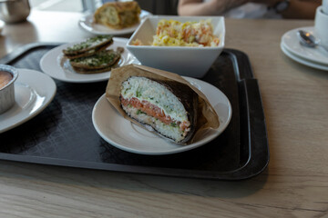 A close-up shot of a salmon sushi sandwich on a white plate. The sandwich is wrapped in brown paper and is sitting on a black tray.