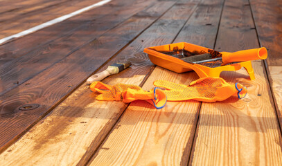 A close-up shot of a partially stained wooden deck with a paint tray, brush, and gloves laid out on...