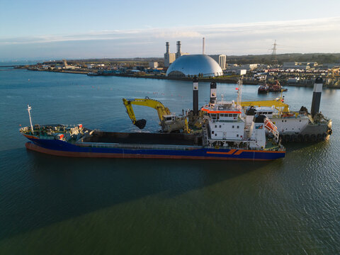 Close-up of backhoe dredger on pontoon and transport barge excavating. Maintenance work at Southampton harbour. Aerial view towards Marchwood Power Station at sunset.