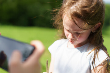 A sad little cute preschooler girl in a green clearing in the forest looks down when she is photographed on a phone. Child loneliness concept