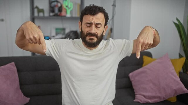 Depressed middle-aged man, wearing a white t-shirt, pointing sadly downwards with fingers, upsetly indicating direction at home. with his gloomy face, he's a pathetic indoor portrait.