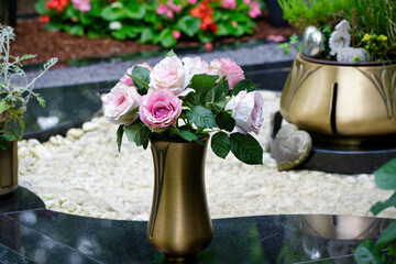 a slender brass vase with an artificial bouquet of roses on a marble grave in a cemetery