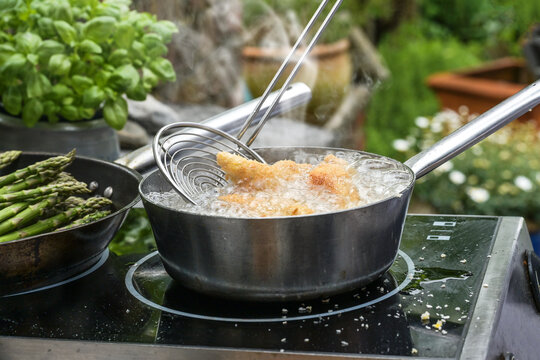 Slotted spoon takes golden deep-fried chicken pieces from a steaming pan with bubbling boiling oil, outdoor kitchen stove surrounded by potted herbs and vegetables, copy space
