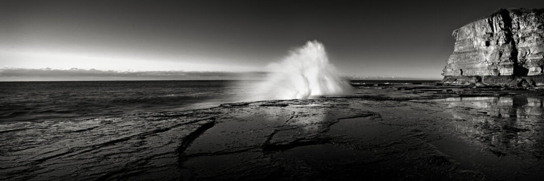 Large ocean wave crashing over rock shelf under cliffs