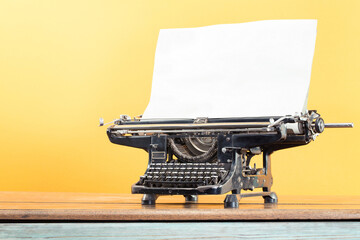Retro typewriter from circa 1930s with sheet of paper blank on wooden desk front yellow wall background. Vintage old style filtered photo