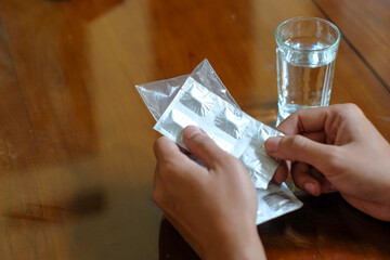 Hand holding a pack of medicine pills with a water glass on wood table background, for medical and healthcare concept.