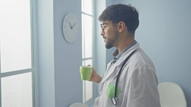 A young bearded man in glasses, wearing a stethoscope and white coat, enjoys a coffee break in a bright hospital room.