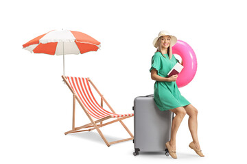 Young woman sitting on a suitcase and holding a passport and a swimming ring next to a beach chair