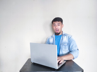 Adult Asian man sitting in front of his laptop showing shocked face expression