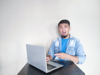 Adult Asian man sitting in front of his laptop showing shocked face expression