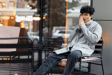 Fashionable man sitting outdoors at a cafe with a cigarette in his mouth and hands on his face