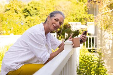 Smiling mature woman holding smartphone and coffee cup, relaxing on porch