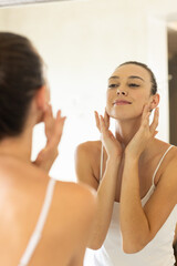 Applying skincare, young woman looking in mirror and touching face at home