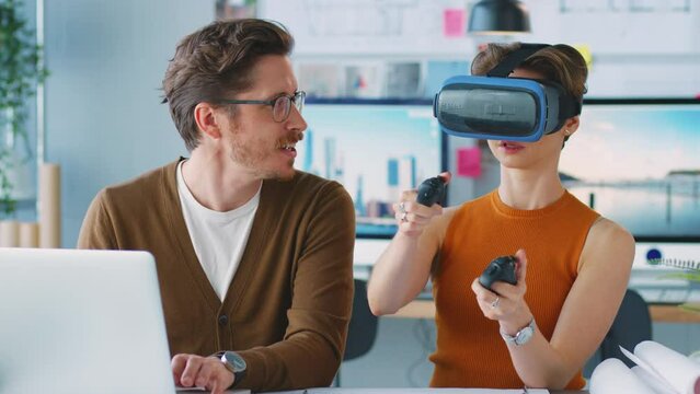 Female architect wearing virtual reality headset in office working next to male colleague at desk with laptop - shot in slow motion