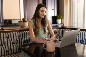 Working from home, young woman using laptop and drinking coffee in kitchen
