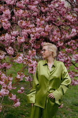 A blonde woman in green outfit poses surrounded by cherry blossoms outdoors