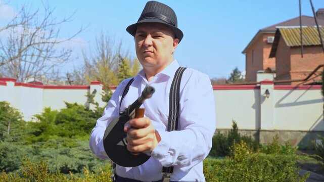 A man in suspenders and a fedora hat aims a vintage tommy gun with determination in a garden setting.