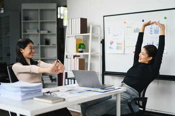 Two Young Women Relaxing and Stretching in a Modern Office During a Break with Laptops and Business Charts in the Background