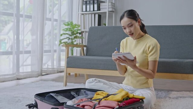 Young woman in casual wear packing a suitcase, checking and writing a list at home. Focus on organization and preparation for a trip