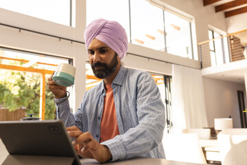 Using tablet and drinking coffee, man working from home in modern living room